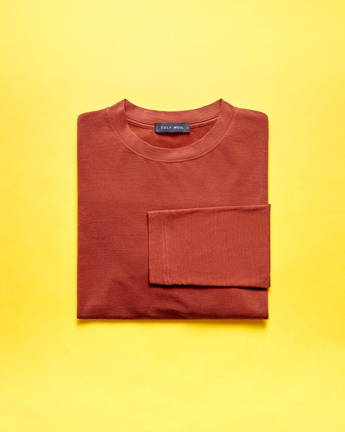 A folded rust-coloured wool long-sleeve t-shirt with a visible neck label against a bright yellow background.