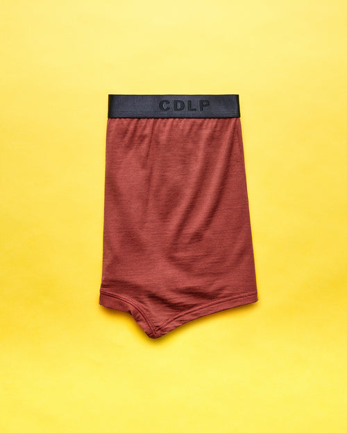 A pair of reddish-brown boxer briefs featuring a black elastic waistband with the CDLP logo, laid flat on a yellow background.