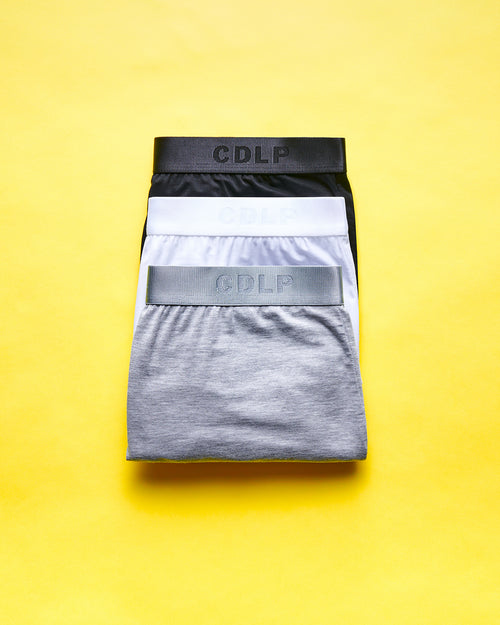 Three folded boxer briefs in black, white, and grey with branded waistbands, stacked on a yellow background.